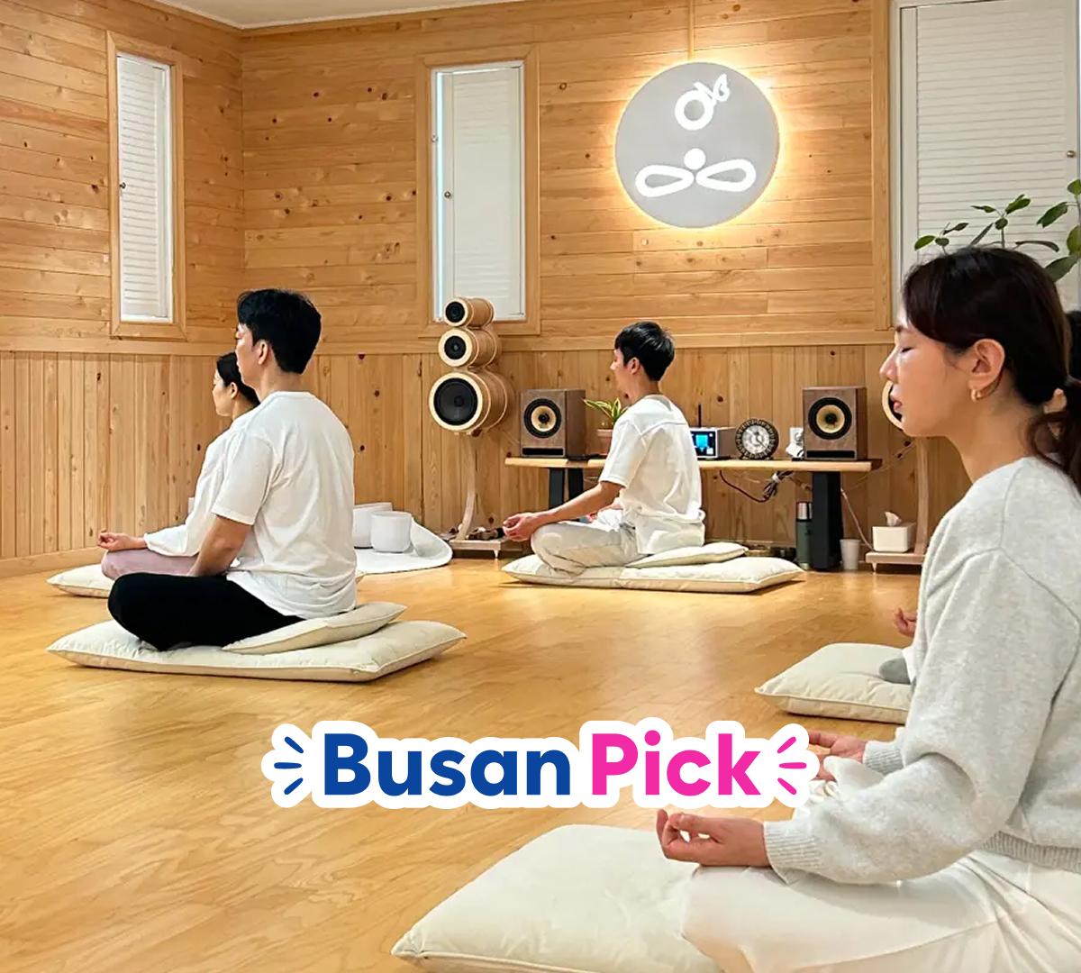 Wood-paneled meditation room with participants seated on floor cushions, playing handpans and crystal singing bowls during sound bath session.