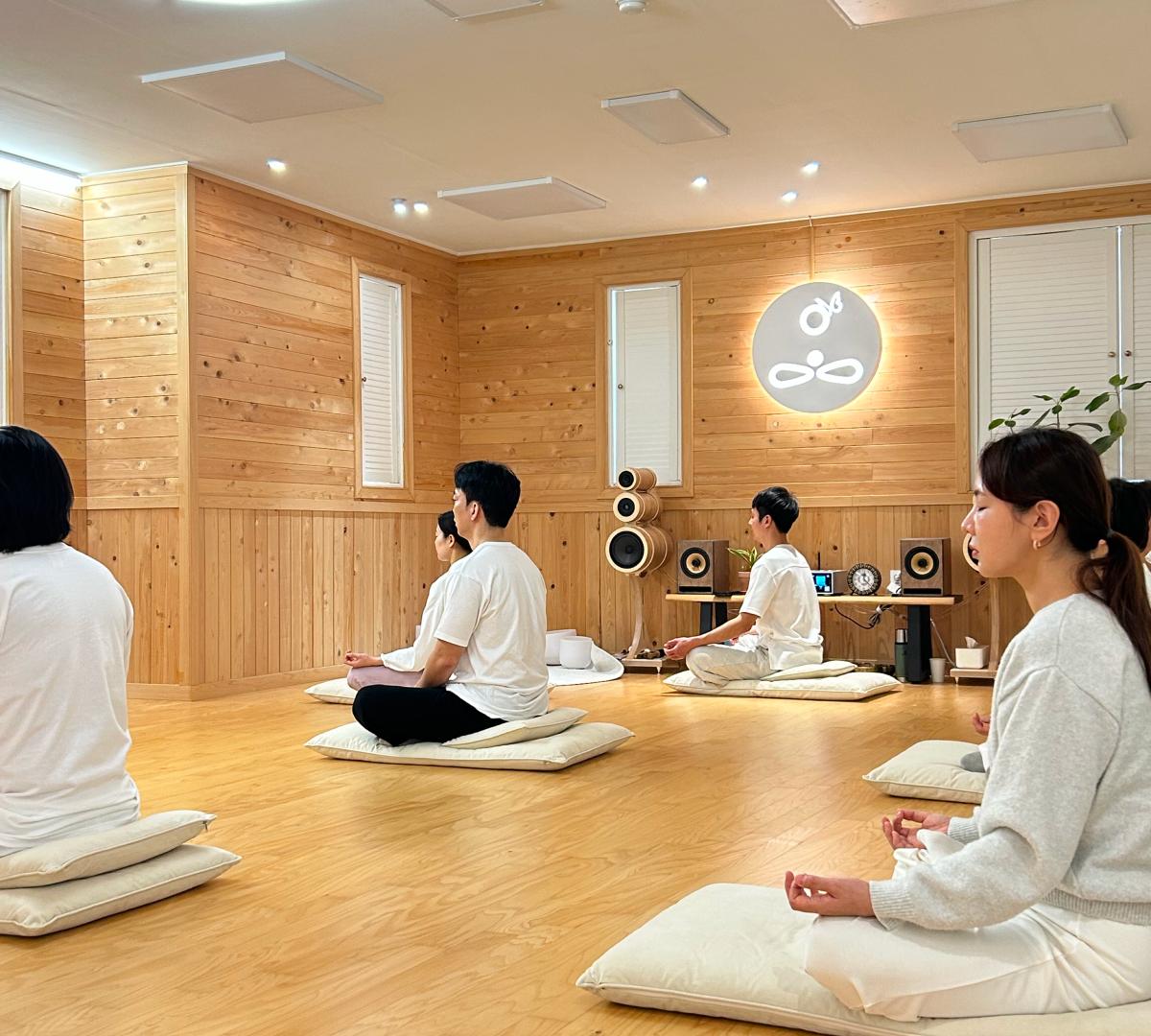 Wood-paneled meditation room with participants seated on floor cushions, playing handpans and crystal singing bowls during sound bath session.