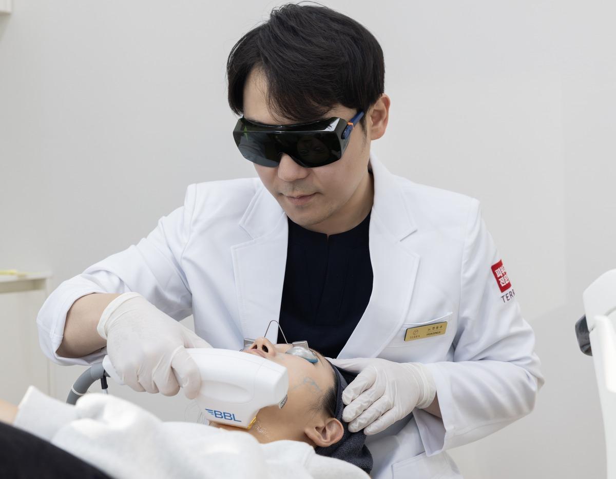Doctor in white coat using handheld BBL device on reclining patient's lower face during clinic treatment