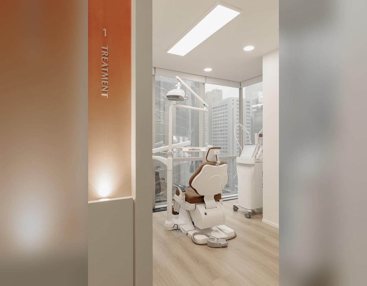 Bright modern treatment room with white dental-style reclining chair, medical equipment and floor-to-ceiling city windows.