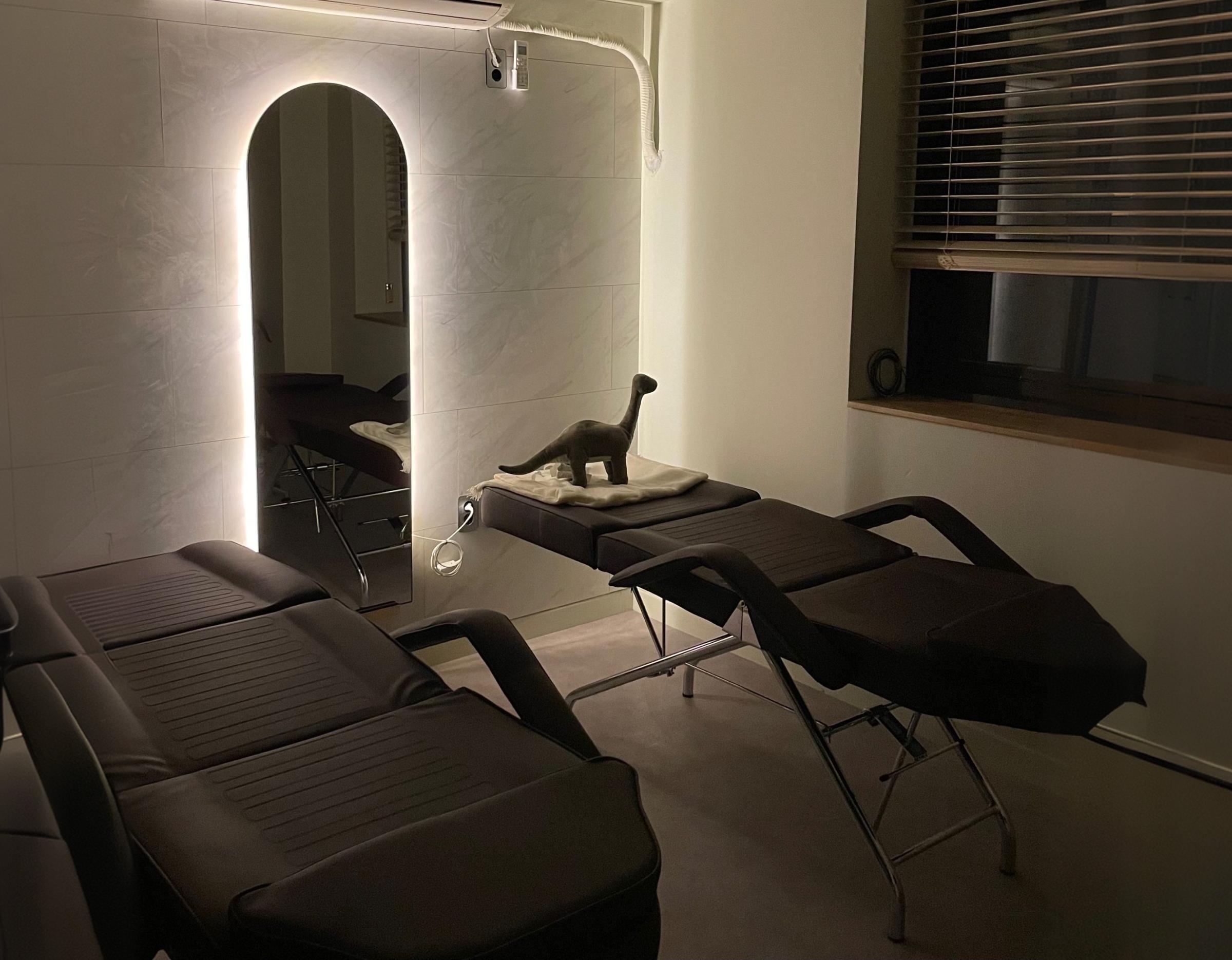 비브로우 홍대 treatment room with two dark reclining salon beds, backlit arched mirror and small dinosaur decor on towel
