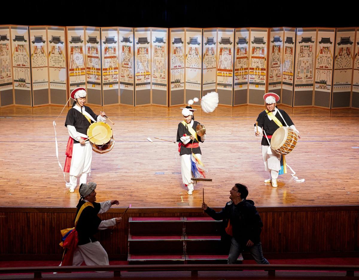 Three Korean traditional musicians on stage in colorful hanbok playing drums and jingles before a painted folding screen backdrop.