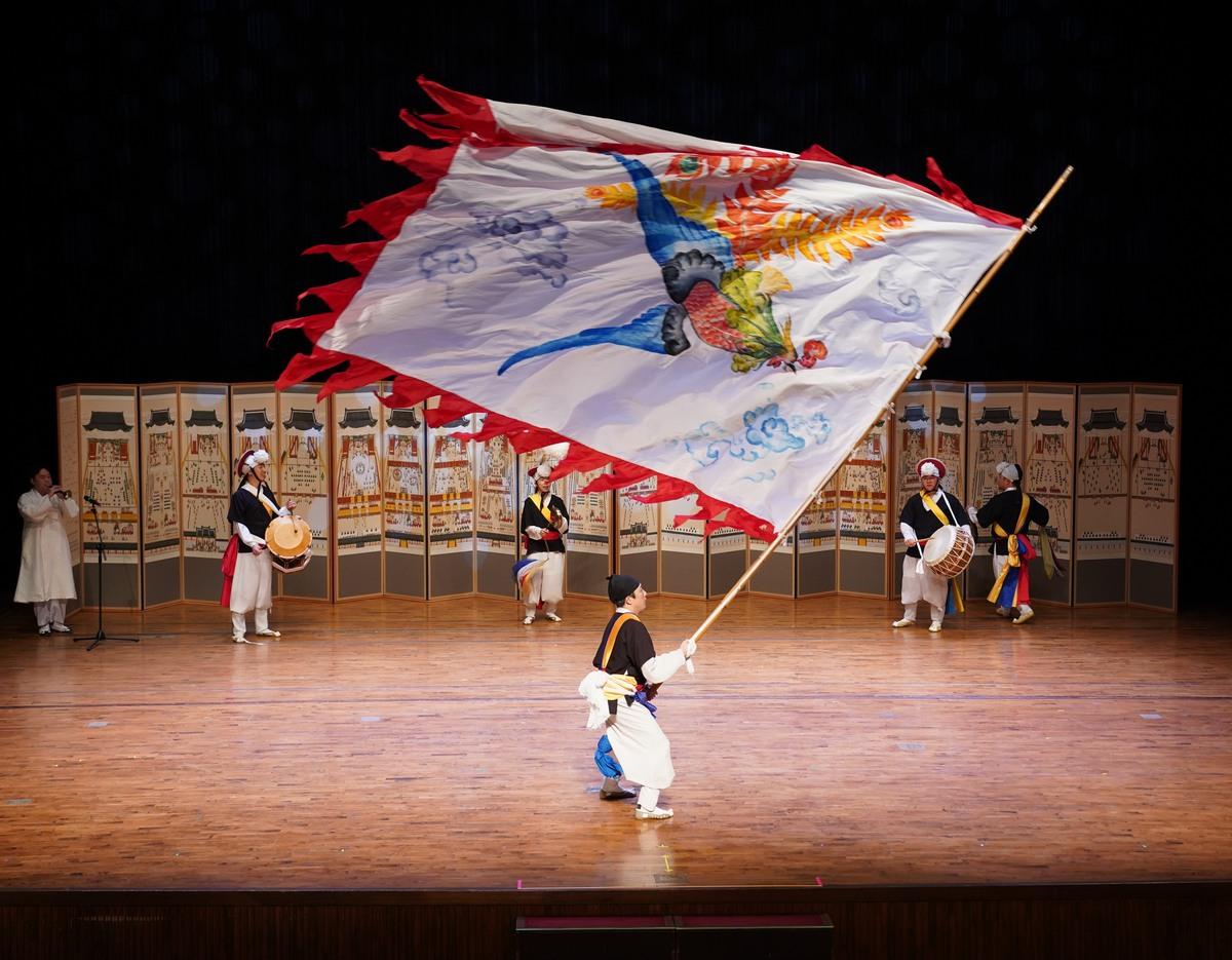 Performer in hanbok twirling a large painted flag with a phoenix onstage, musicians and painted folding screens in background