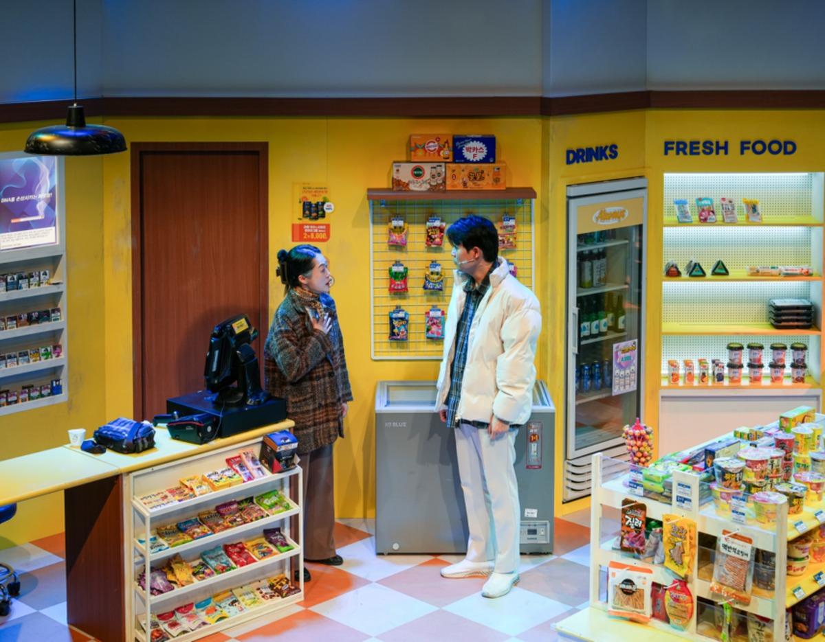 Stage set of a brightly lit yellow convenience store with two actors talking by a freezer, shelves stocked with snacks and drinks.