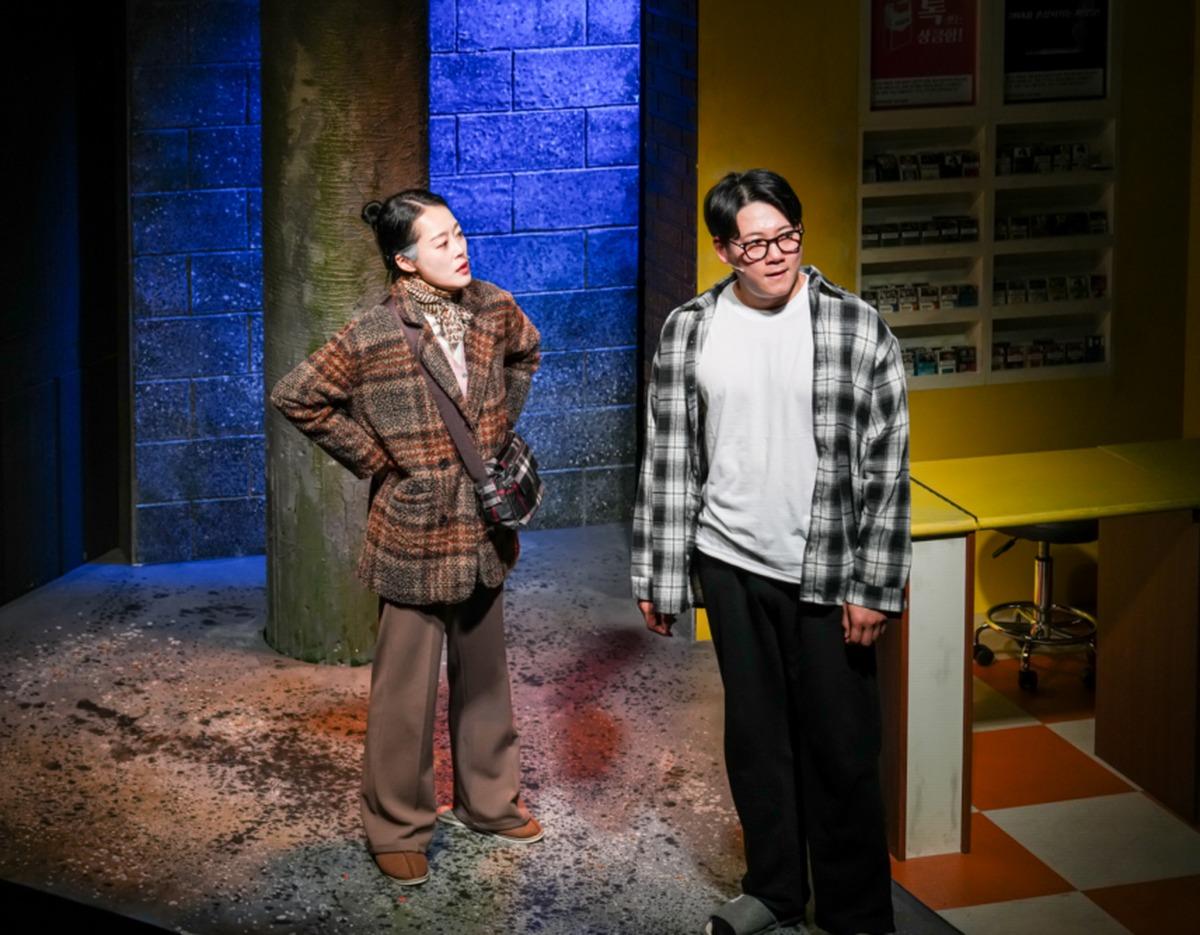 Two actors on a dimly lit stage set of a convenience store; one in a plaid coat and crossbody bag, the other in a checkered shirt.