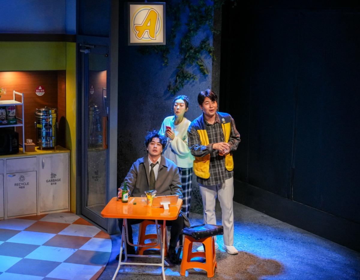 Stage scene from Seoul Music Drama ticket showing three actors in a convenience store set, one seated at orange table with drinks and two standing nearby.