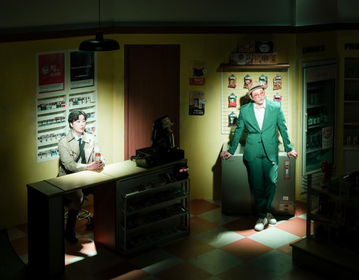 Dimly lit convenience store stage set with cashier at counter and man in green suit leaning by fridge under spotlight