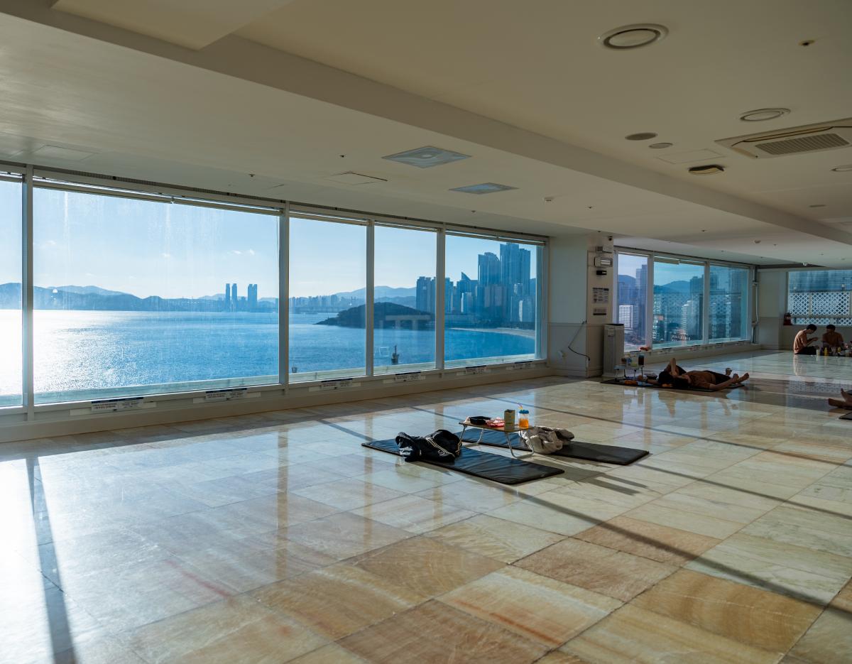 Spacious healing room with mats and belongings on tiled floor, floor-to-ceiling windows showing Haeundae ocean and city view
