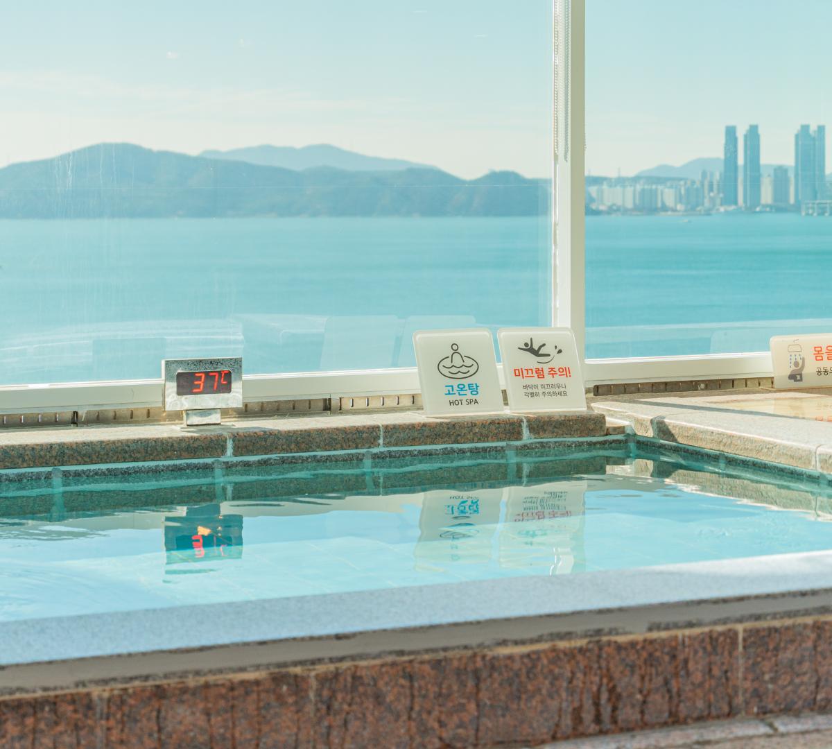 Indoor hot spa pool by a large window with a 37°C digital thermometer and ocean view of Haeundae and distant skyline