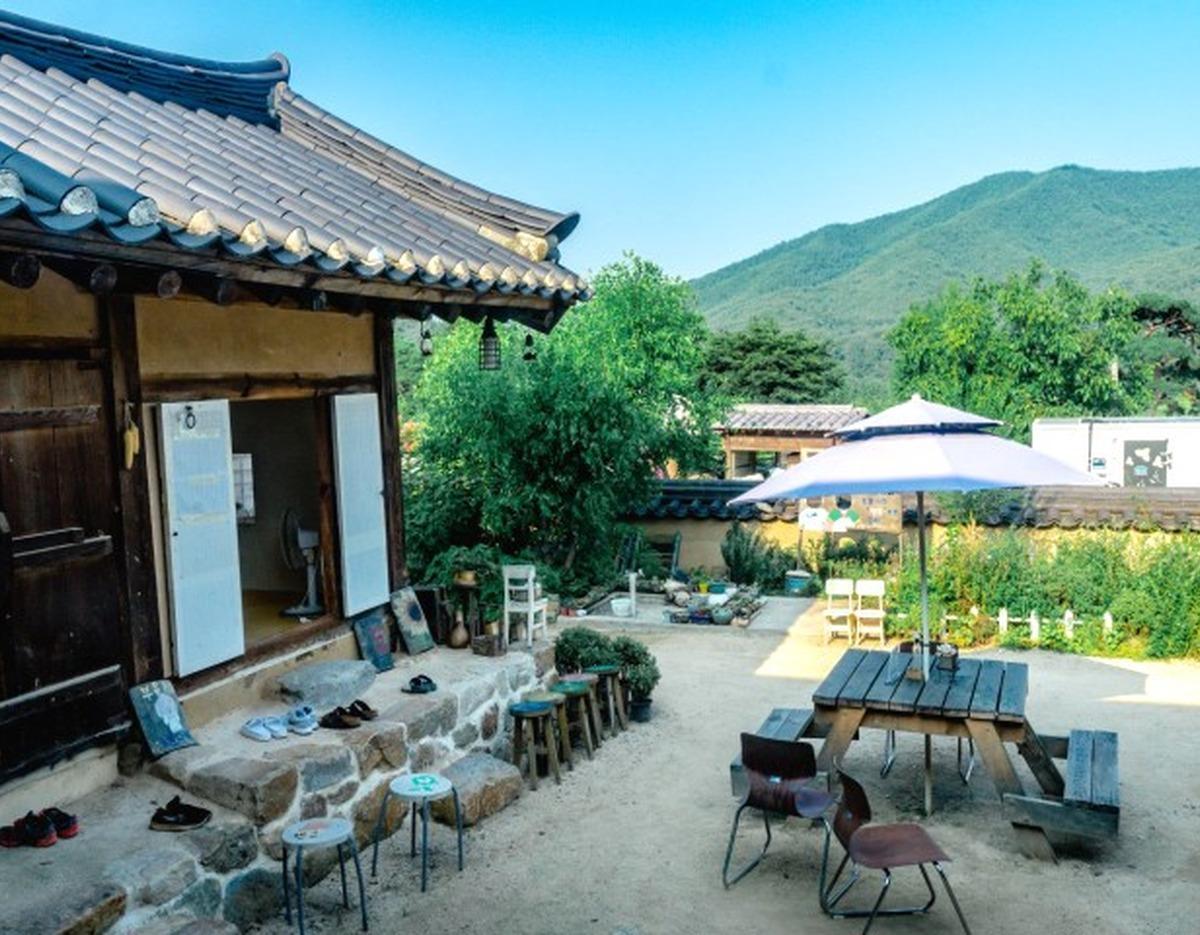 Traditional Korean hanok courtyard with low stone steps, stools and shoes, picnic table and umbrella, green mountain backdrop