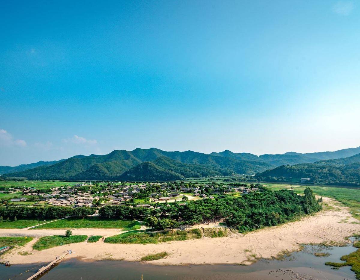 Panoramic view of Andong Hahoe Village with traditional thatched houses by riverbank, sandy shore and green mountains behind