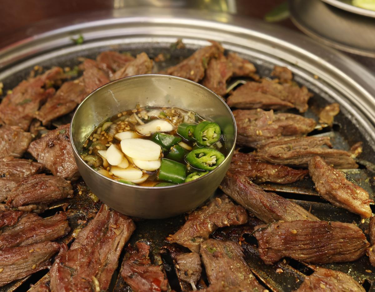 Grilled marinated beef short ribs arranged on a round charcoal grill with metal bowl of soy sauce, sliced garlic and green chili.