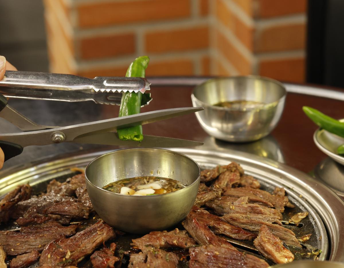 Grilled marinated beef short ribs on a tabletop charcoal grill with dipping sauce bowl and scissors cutting a green pepper
