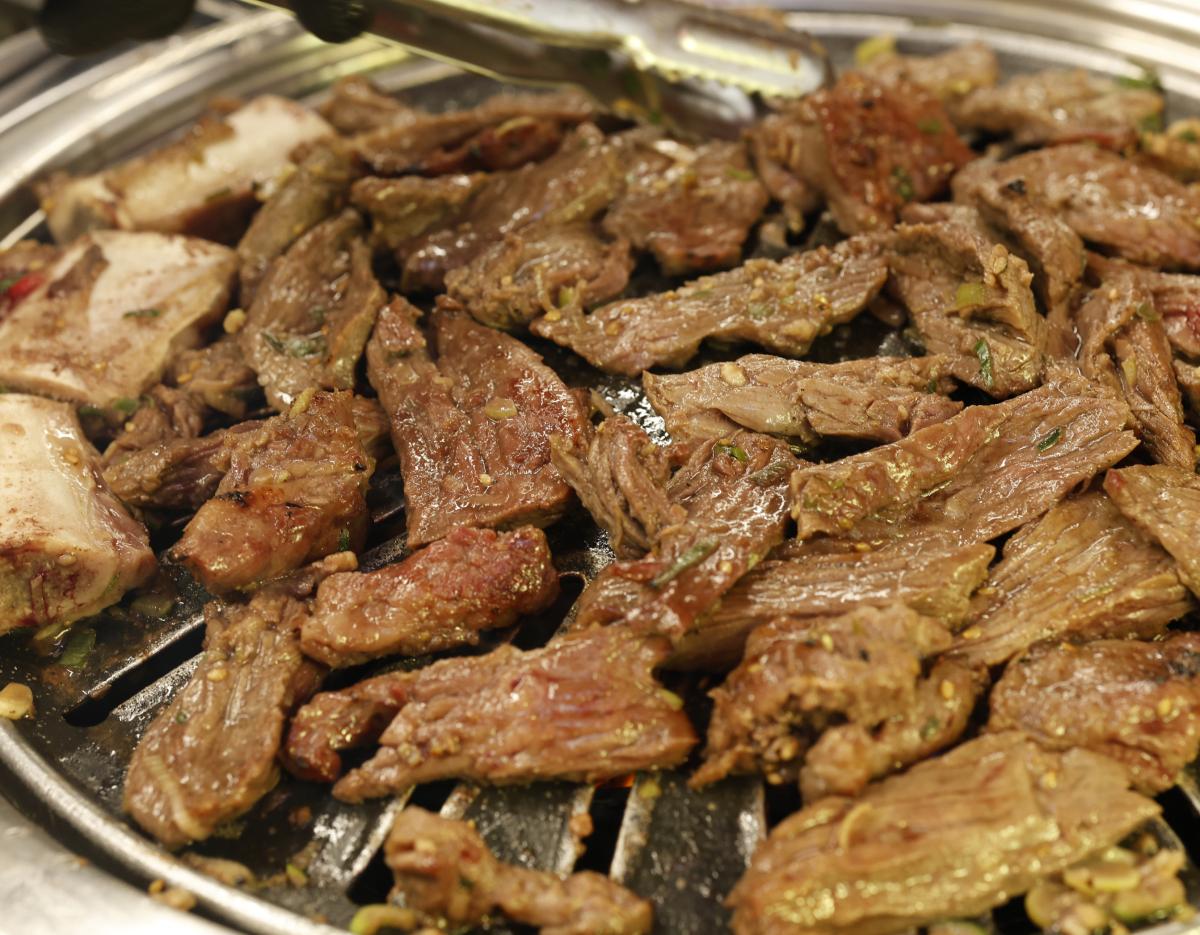 Grilled marinated beef short ribs arranged on a round charcoal grill with metal bowl of soy sauce, sliced garlic and green chili.