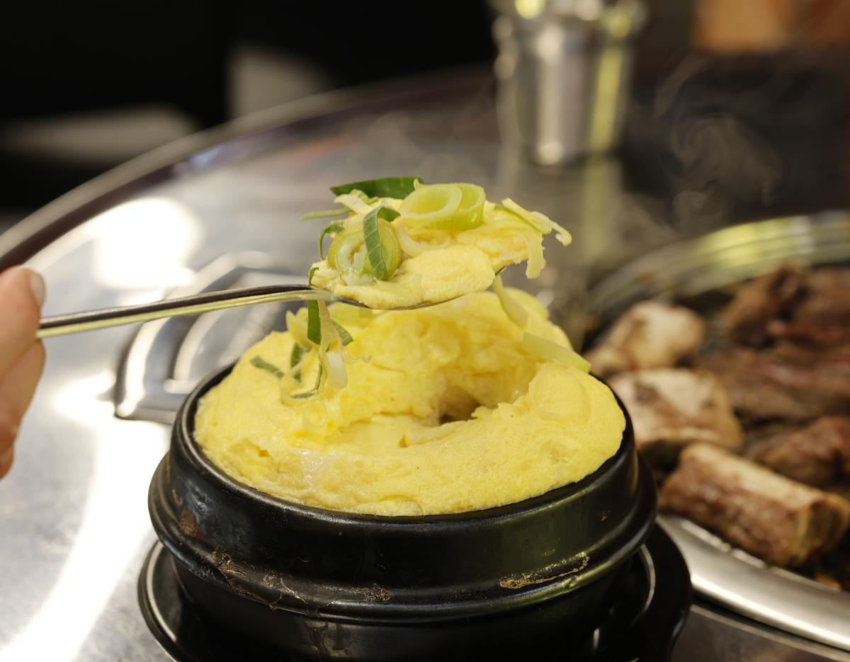 steaming Korean gyeran-jjim (savory steamed egg) topped with sliced scallions in a black stone bowl beside grilled short ribs