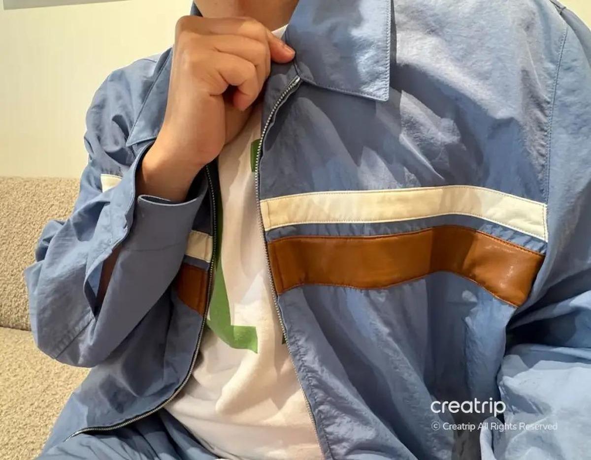 Model wearing a light blue zip-up jacket with white and brown horizontal stripes over a white tee, seated on a beige sofa