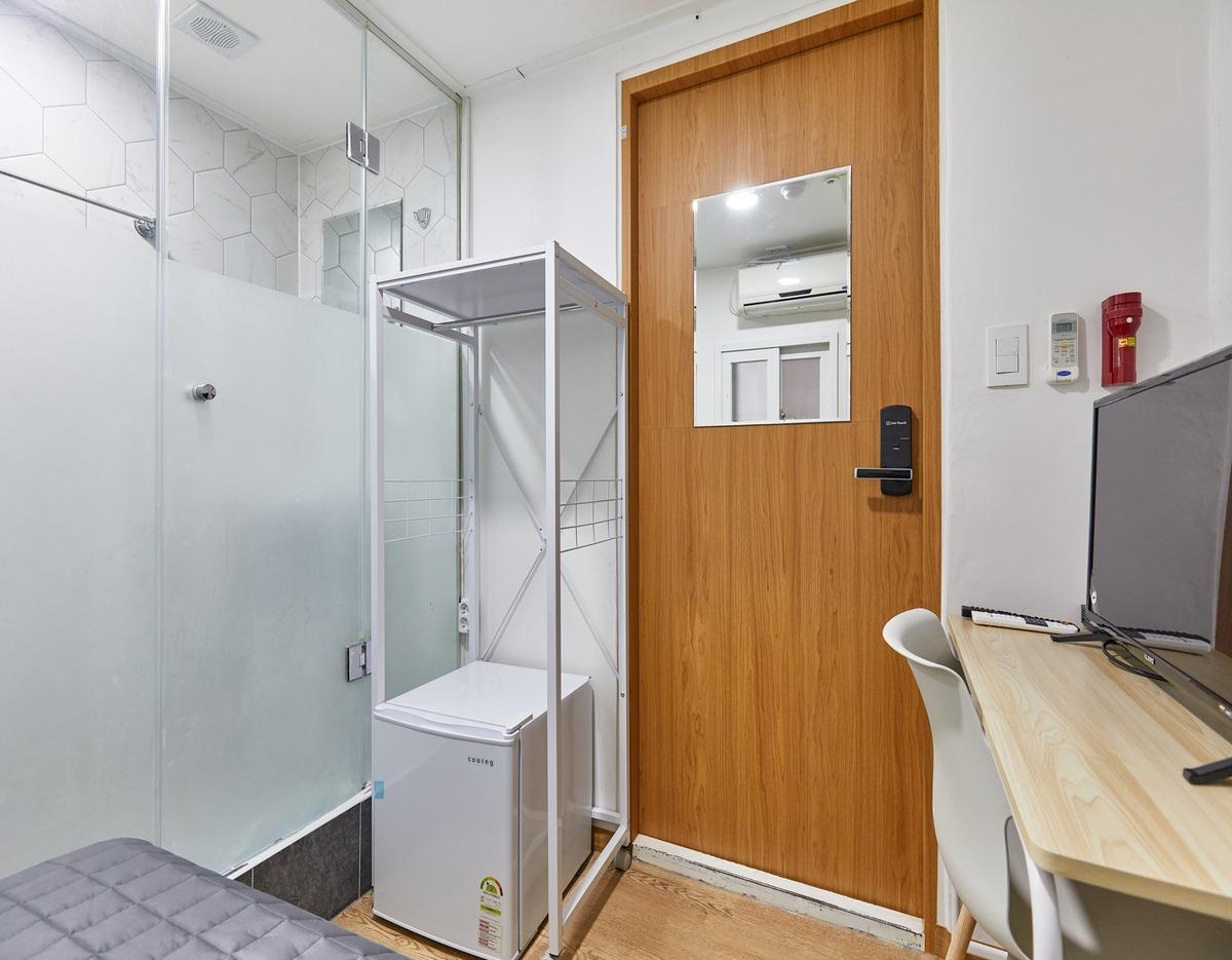 Compact single room with wooden door, small fridge in metal shelving, glass-enclosed bathroom and desk with chair and TV