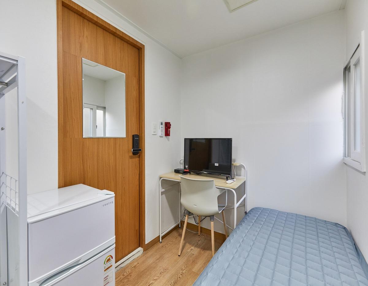 Compact single-occupancy room with a single bed, small desk and chair with monitor, mini fridge, and wooden door window.