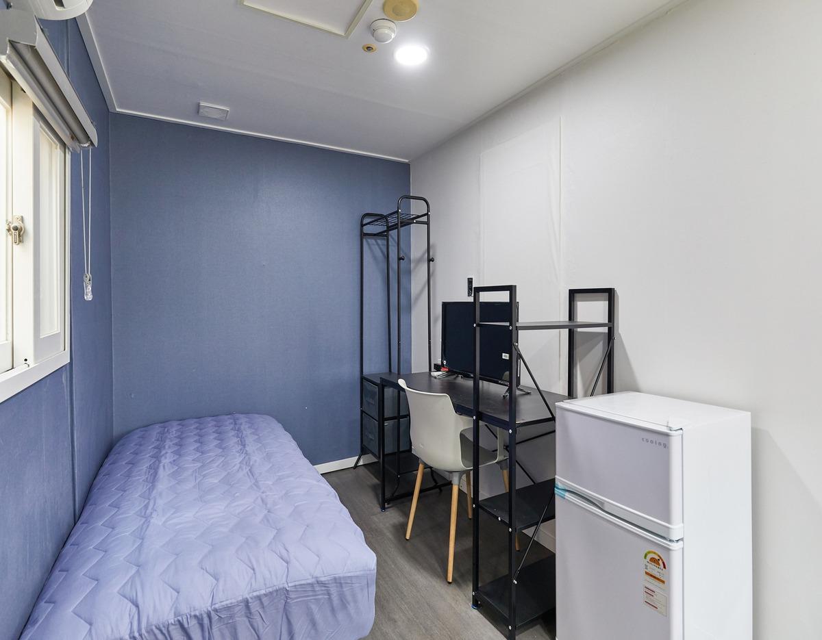 Compact single room with a single bed, desk with monitor and chair, open shelving rack, and a small white refrigerator