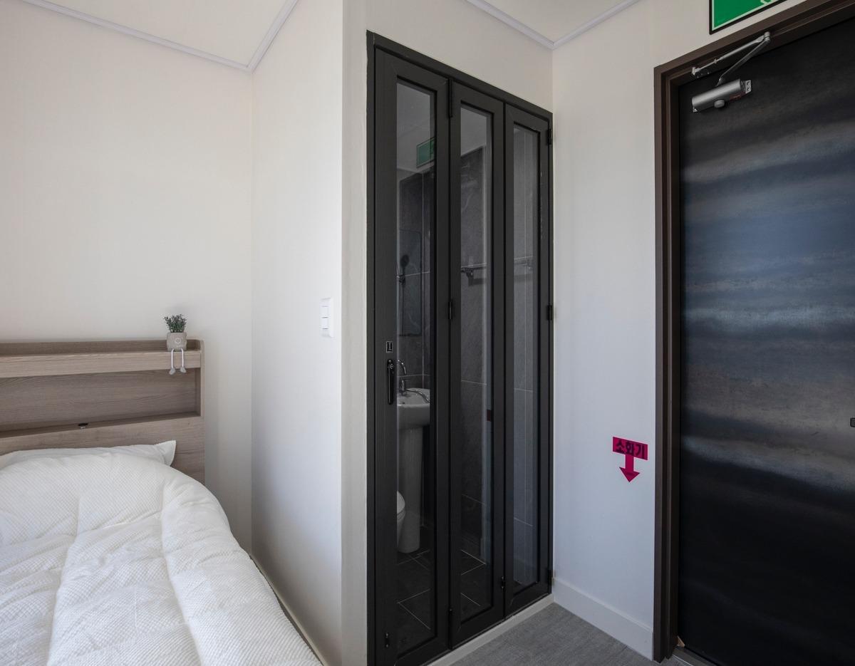 Compact bedroom corner at 숲속휴식 광운대점 showing single bed, small headboard plant, folding glass bathroom door and entrance door.