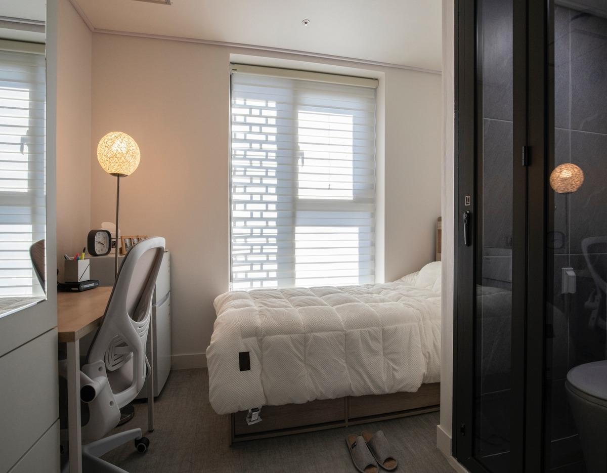 Compact single room with white quilted bed, desk and ergonomic chair by window, lamp and sliding glass bathroom door.