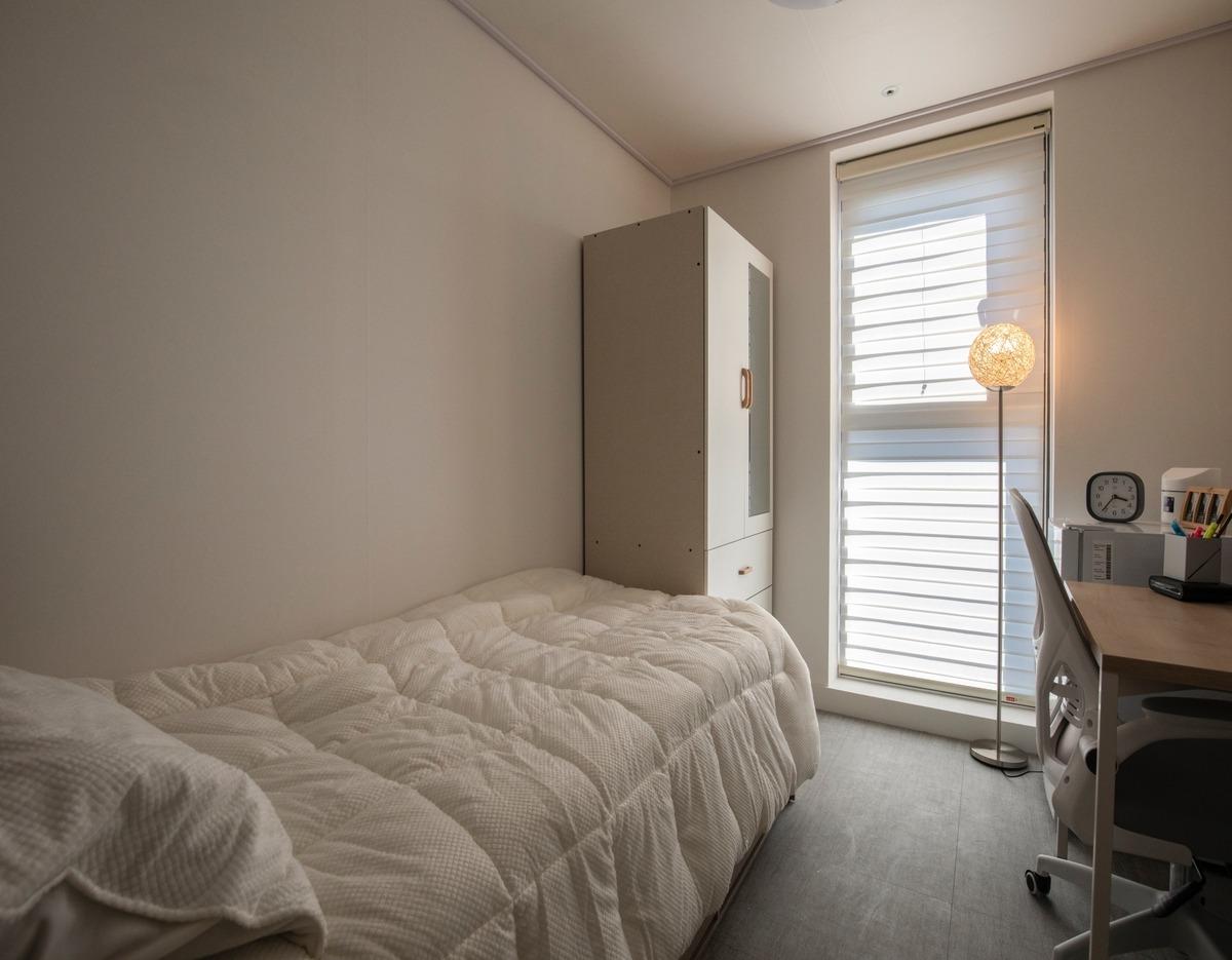 Small single bedroom with white quilted bed, tall wardrobe, desk with chair and floor lamp by a window with blinds