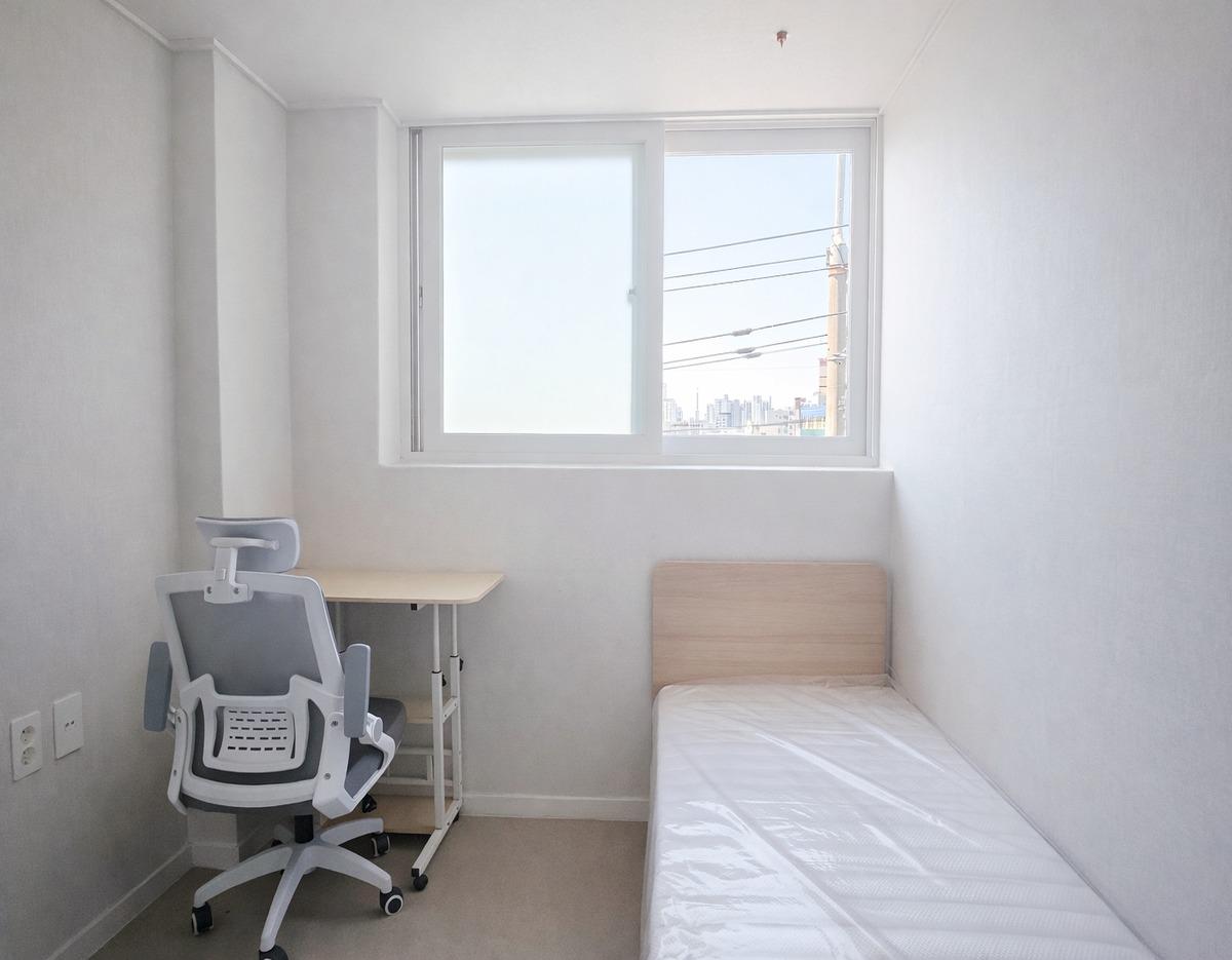 Minimal single room at HUESIK DAERIM with narrow bed, wooden headboard, small desk and ergonomic chair under a window.