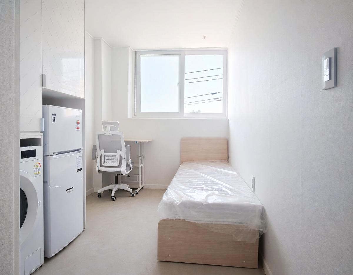 Compact single room with a single bed, desk and ergonomic chair beside a window, plus fridge and washer in white minimalist layout
