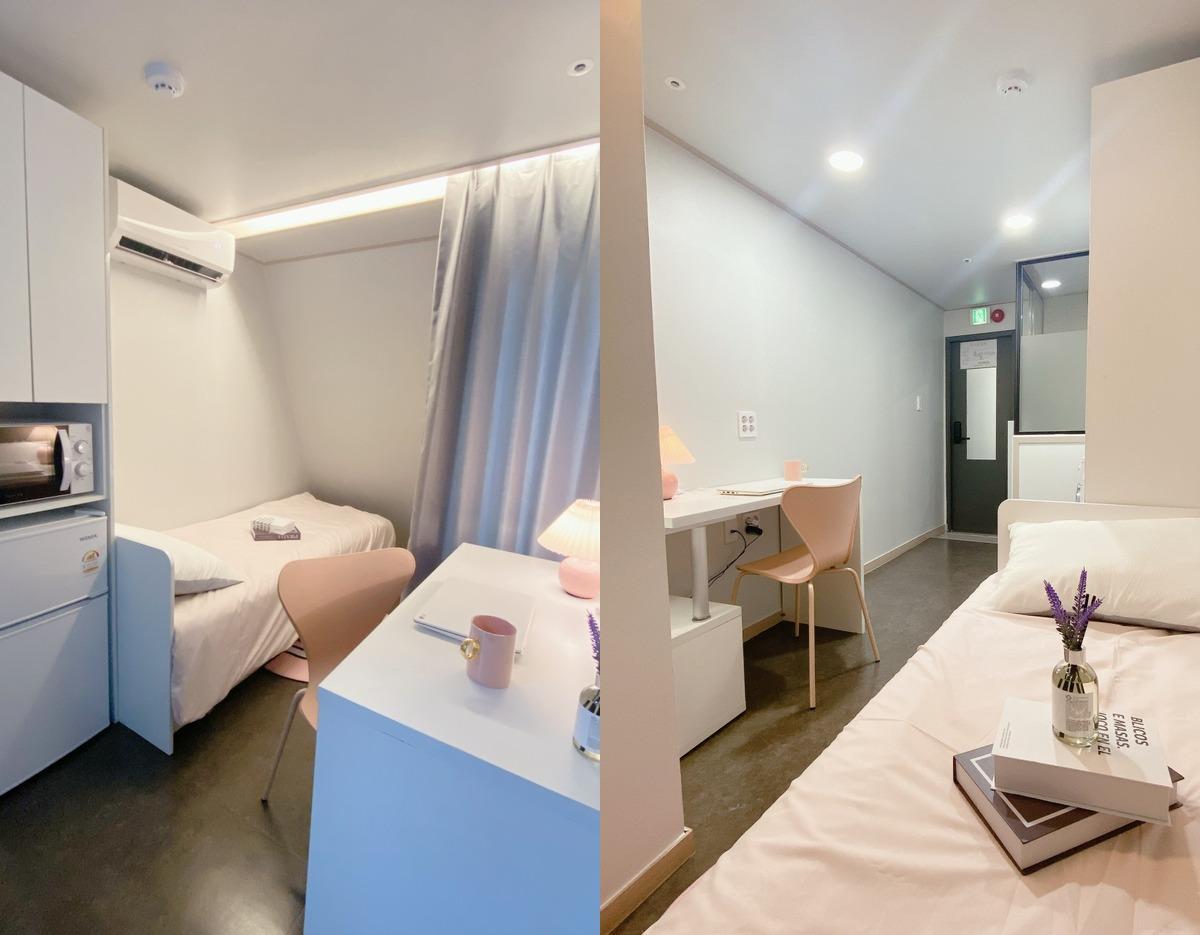 Compact studio room with single bed, desk, chair, small fridge, microwave, air conditioner and soft pink accents
