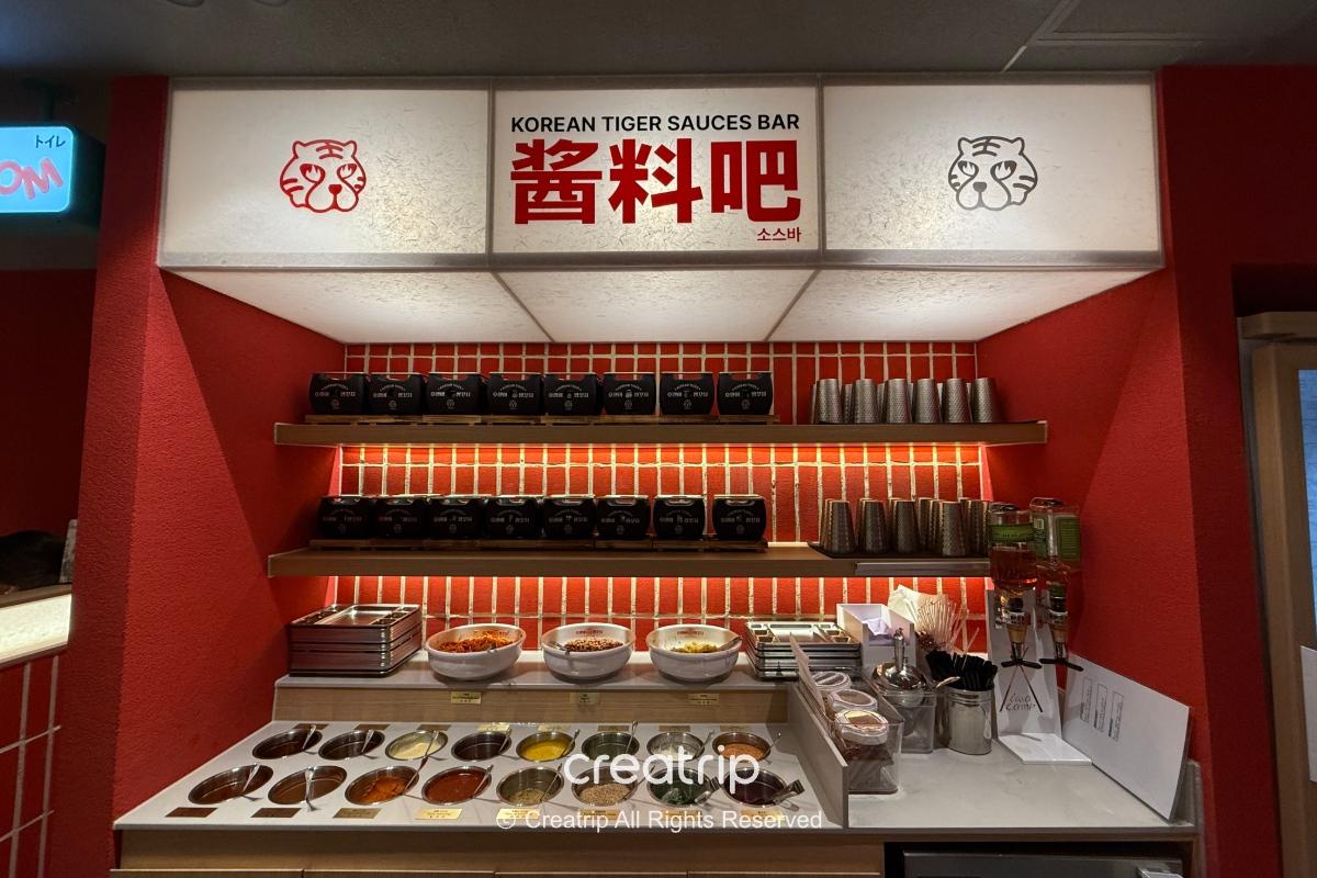 호랭이양꼬치 sauce station with labeled bowls, metal cups, condiments and dispensers against red tiled wall under "Korean Tiger Sauces Bar" sign