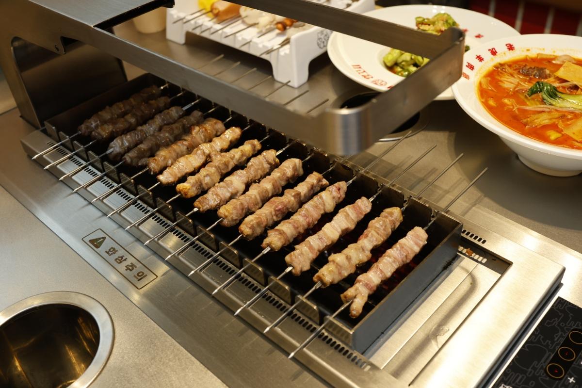 호랭이양꼬치 skewers of tender lamb belly grilling on a smokeless tabletop griddle with side bowls of mala soup and vegetables