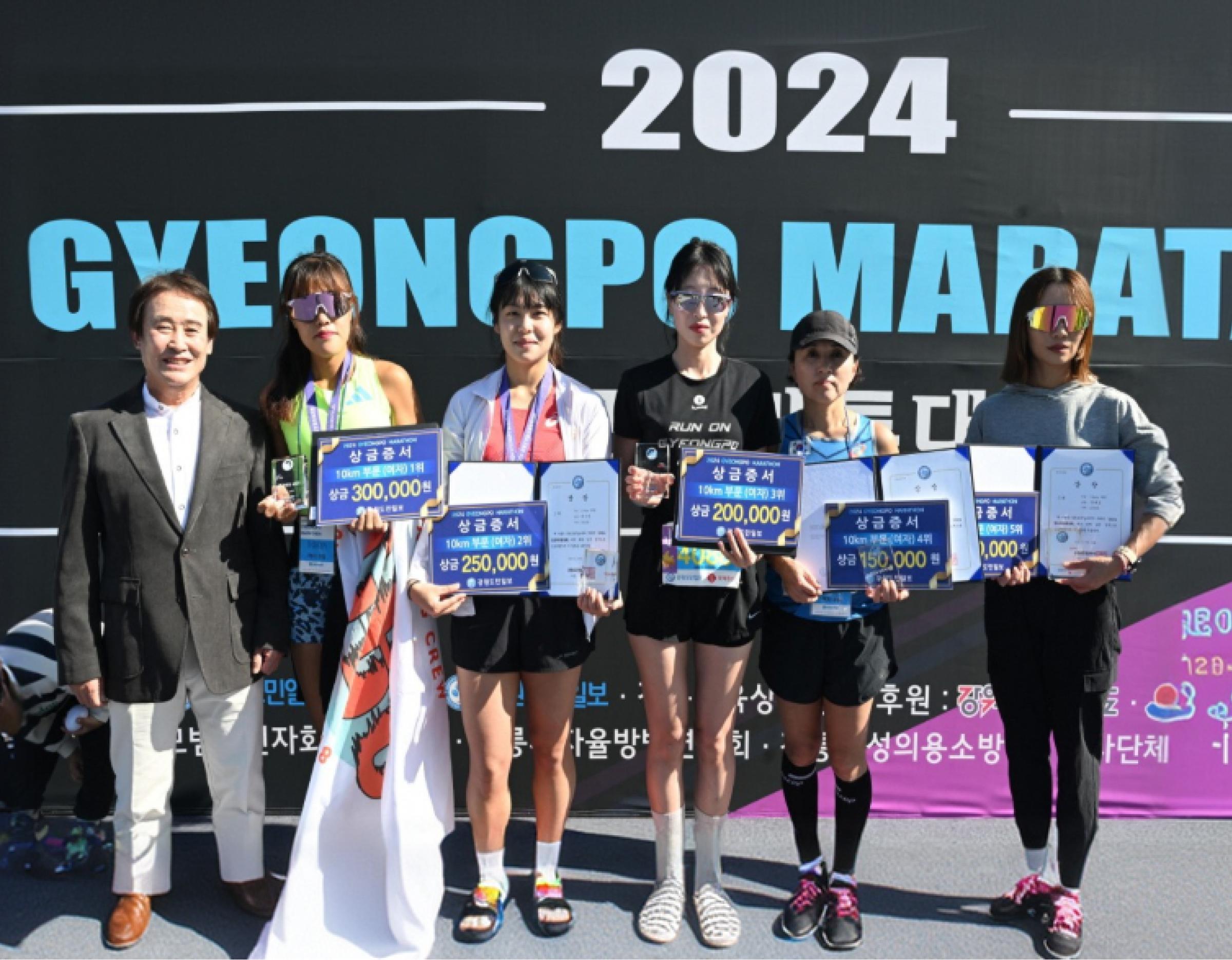 Six people standing on a stage holding placards, trophies and certificates in front of a large "2024 GYEONGPO MARATON" backdrop.