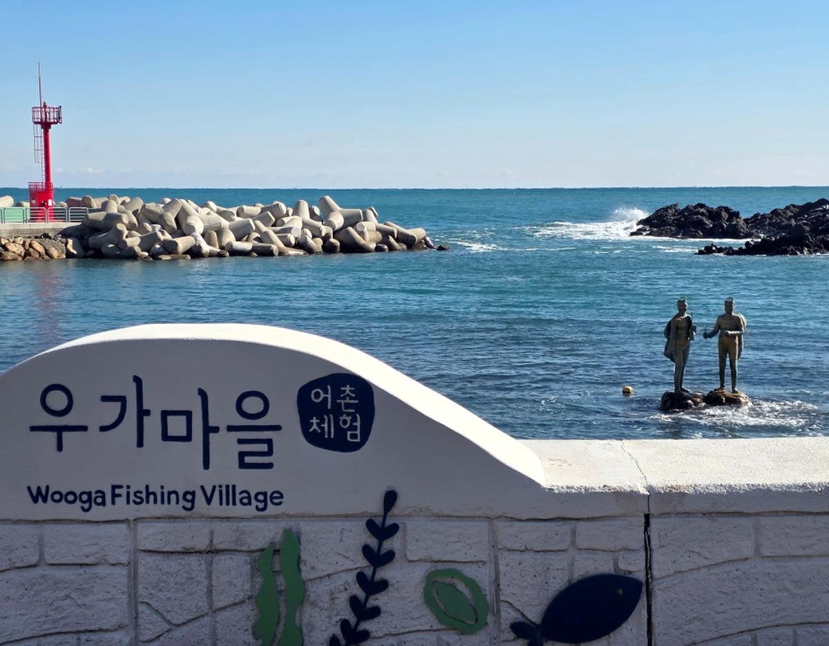 Wooga Fishing Village sign on seawall with blue ocean, tetrapod breakwater, red lighthouse and twin statues on rocks in water