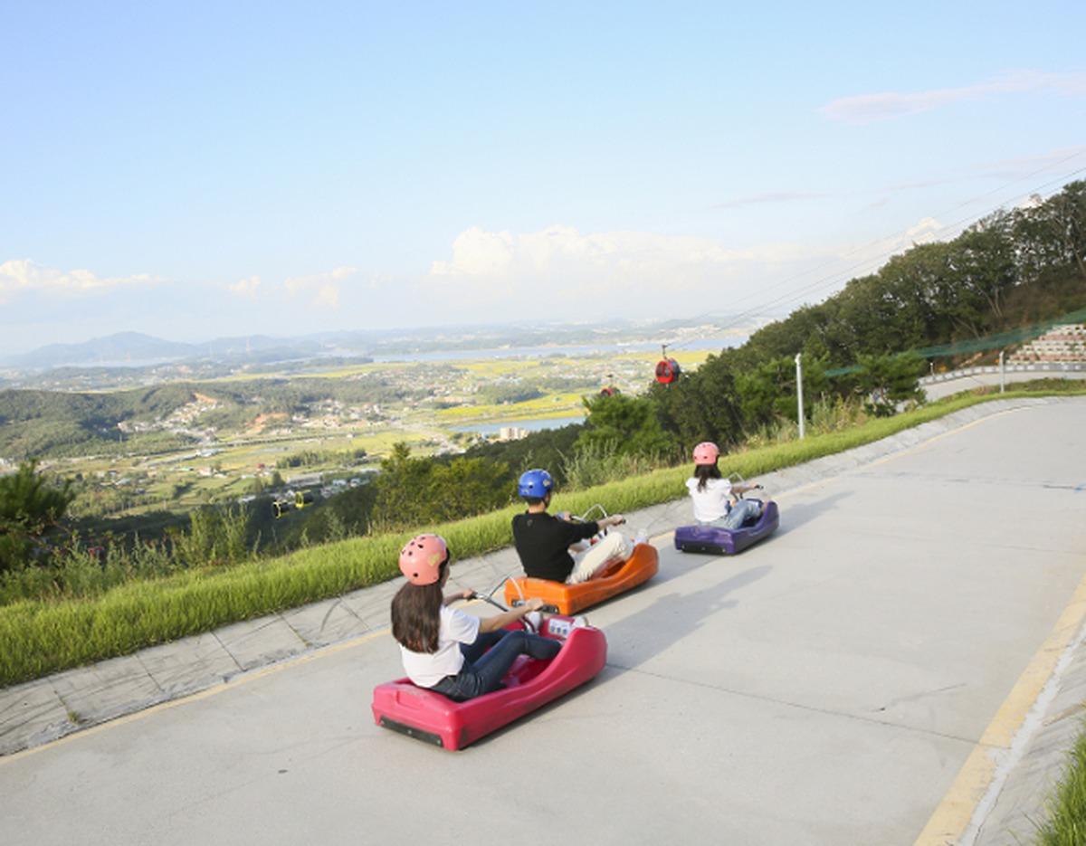 Three riders wearing helmets on colorful luge carts descending a paved track with green hills and valley views behind them