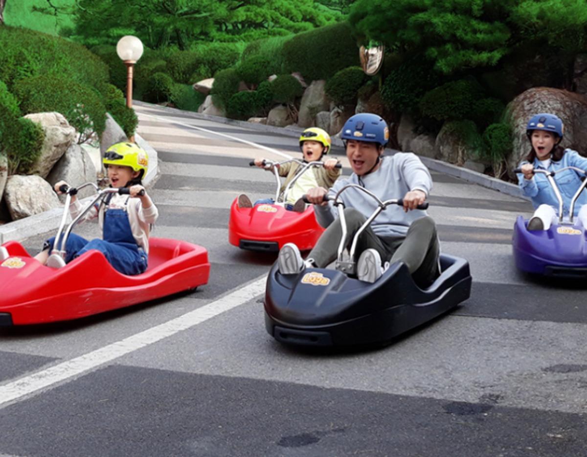 Four people wearing helmets ride colorful downhill luge carts on a paved track lined with rocks and trimmed greenery.