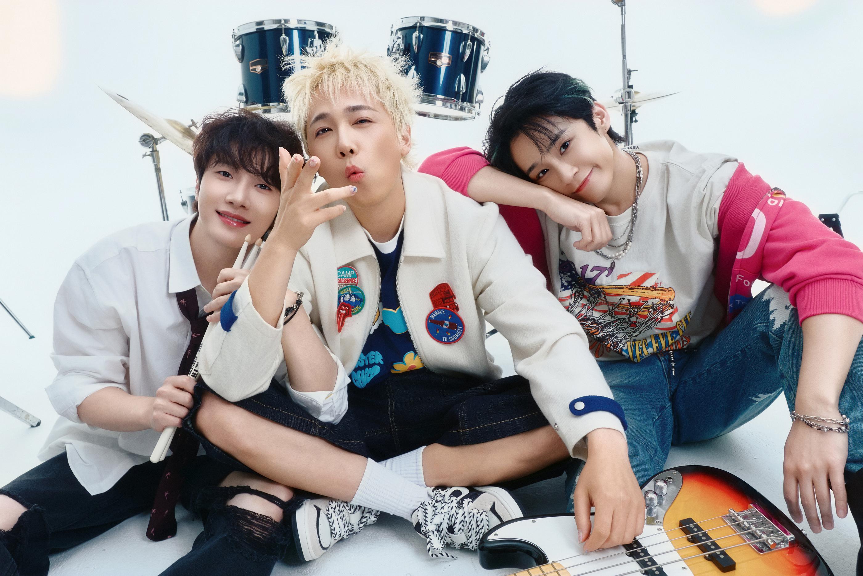 three young performers sitting cross-legged on studio floor with drum kit and electric bass, casual festival-style outfits