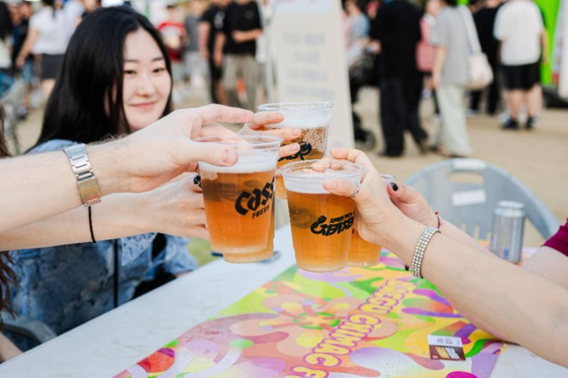 Four hands clinking plastic cups of beer over a colorful festival table at the Daegu Chi-Maek (chicken & beer) festival.
