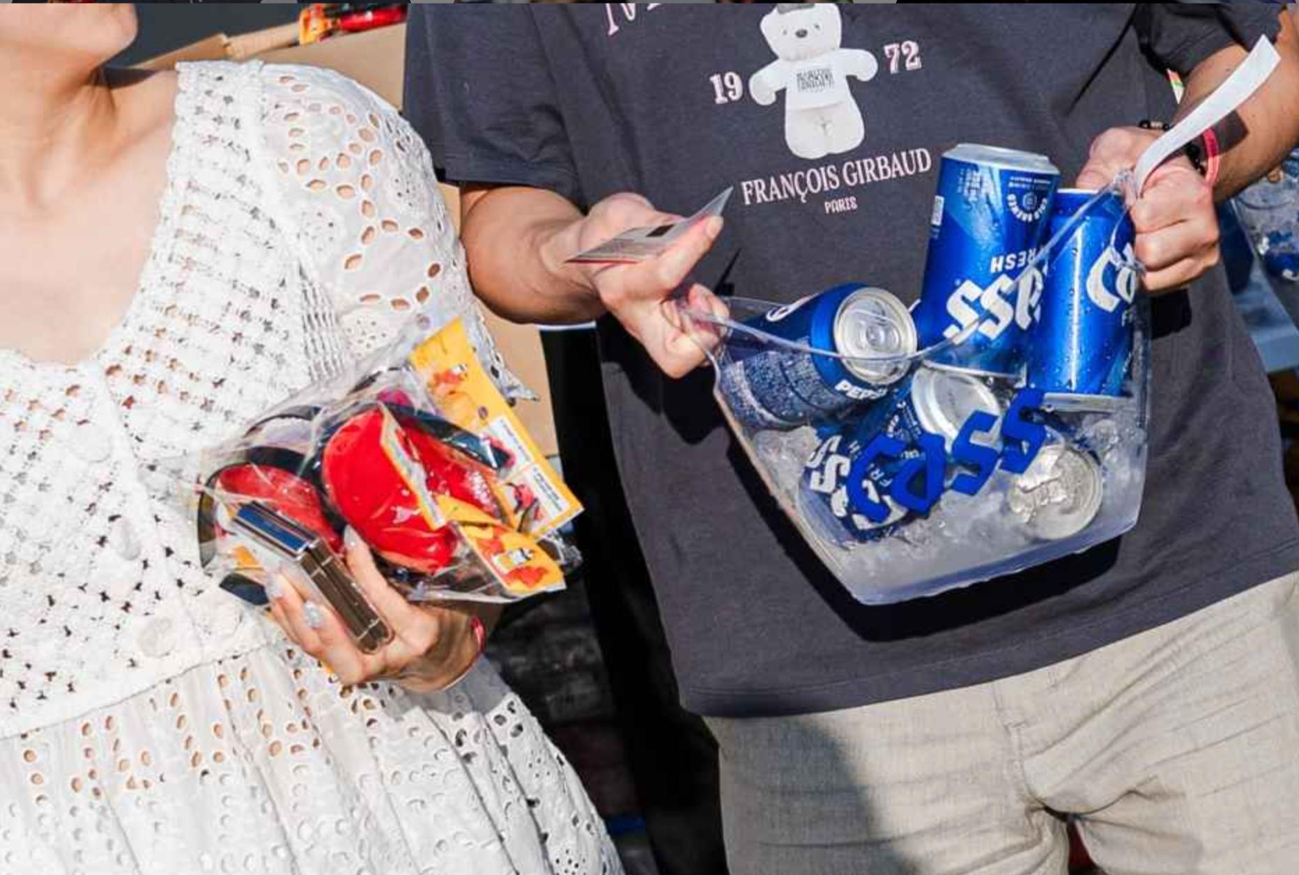 Festival-goers holding plastic trays: one with several canned Asse beer on ice, another with packaged festival toys and a phone.