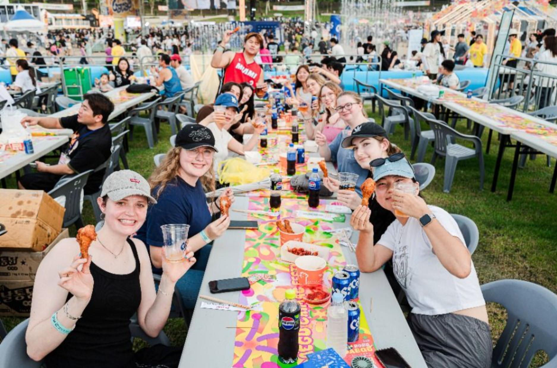 Long table at Daegu 치맥 페스티벌 with people holding fried chicken drumsticks, drinks, colorful table runner and festival crowd in background