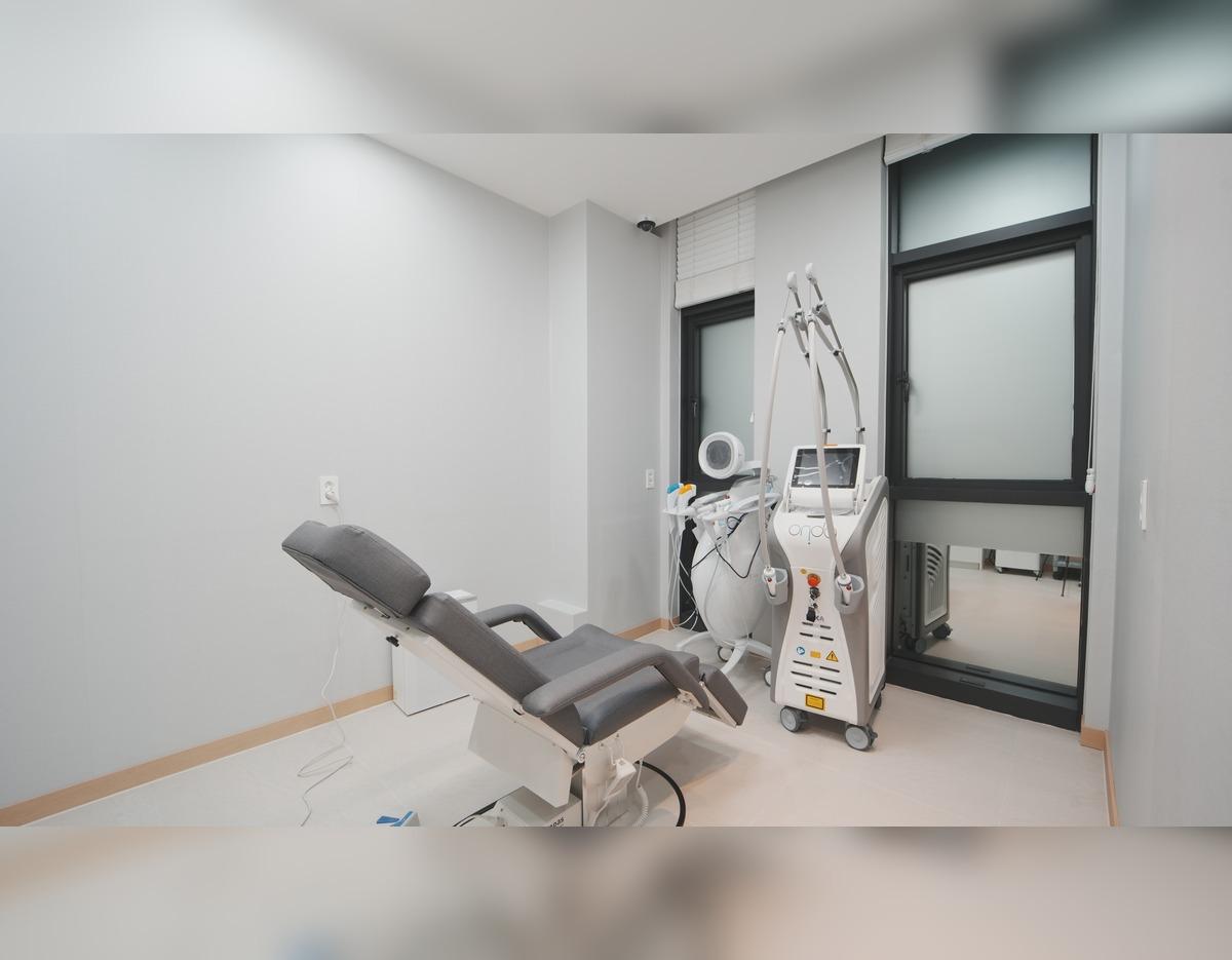 Clinic treatment room with reclining gray exam chair and laser/skin booster machines by window