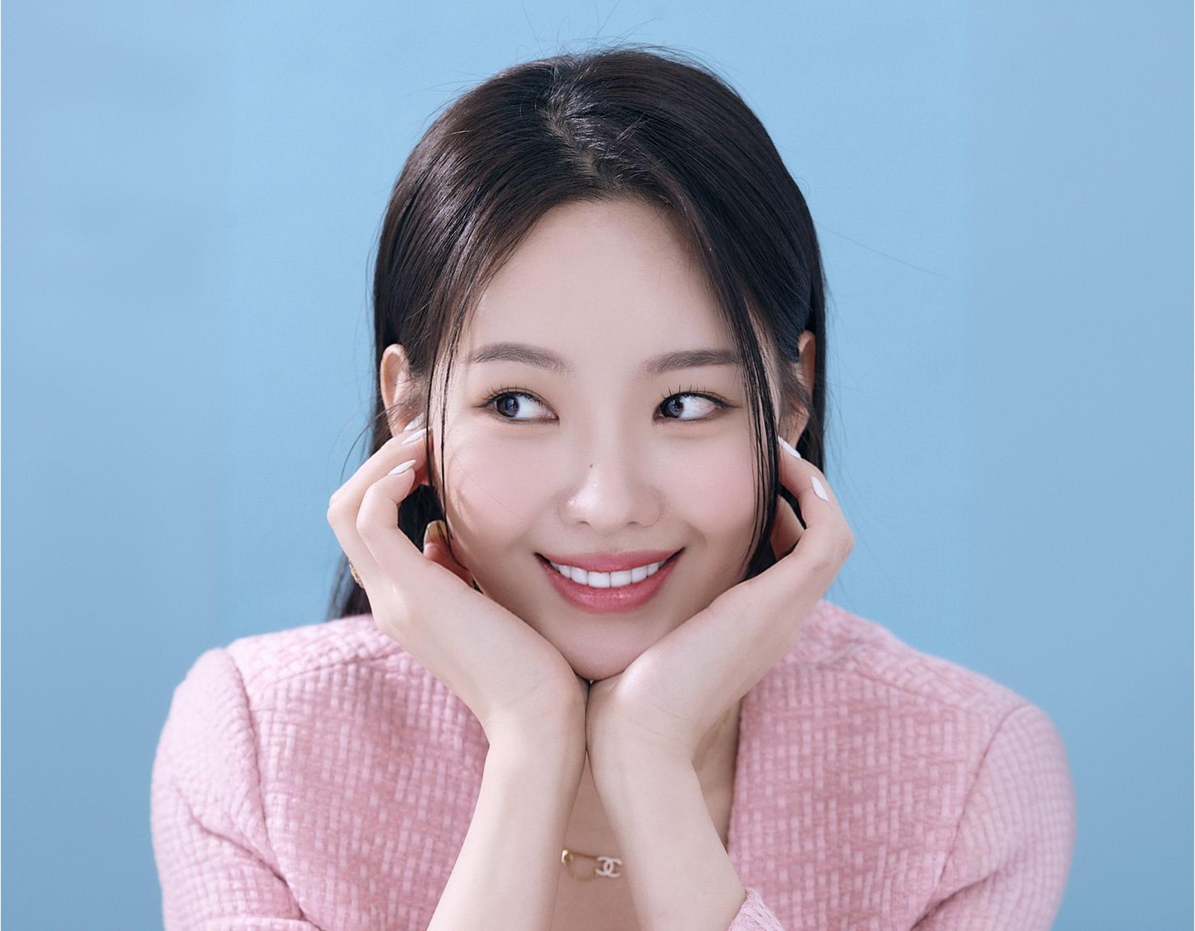 woman in pink textured jacket resting chin on hands against pale blue background, close-up portrait, showing a clean and confident smile after Lucinate laminate dental treatment