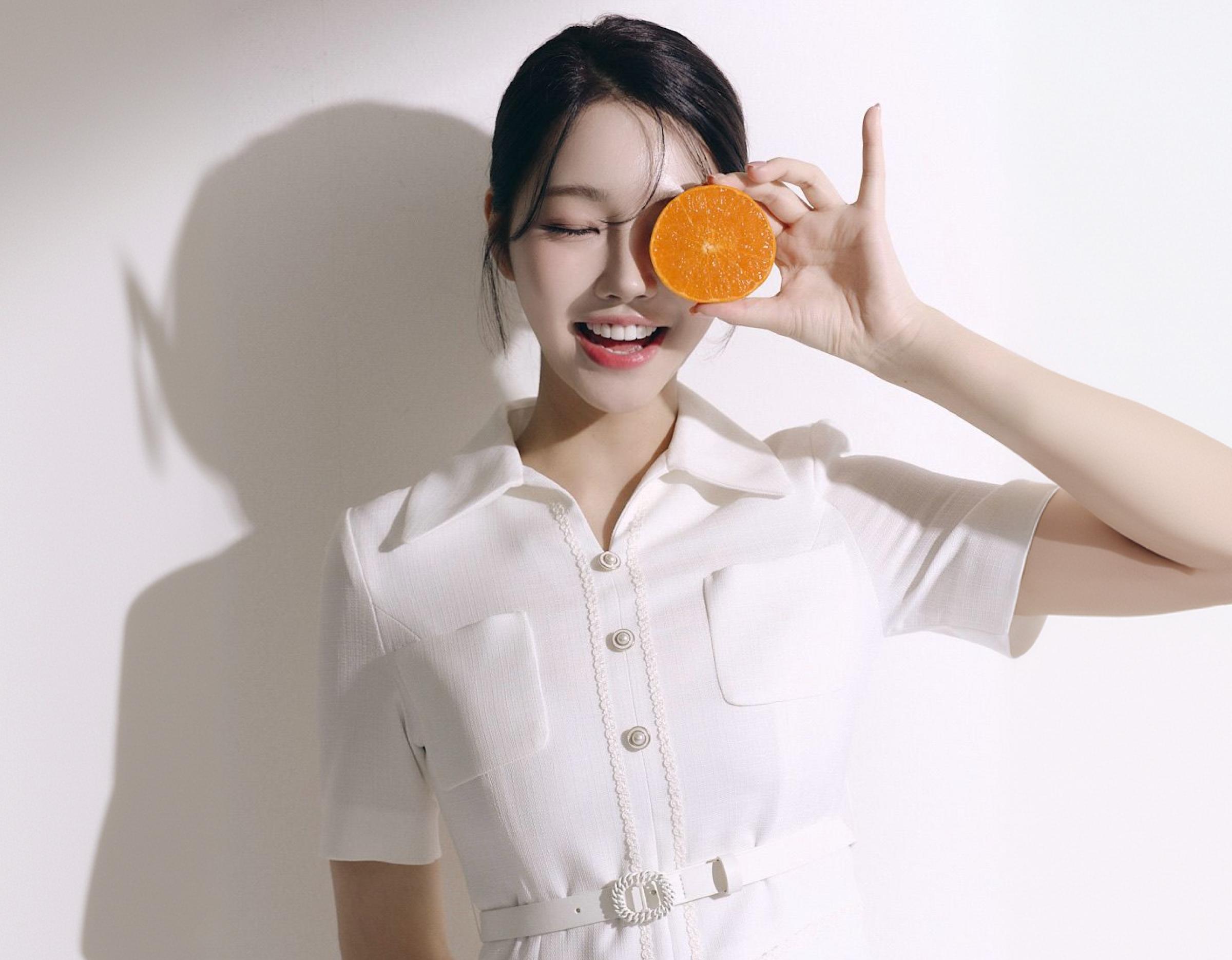 woman in white collared dress holding an orange half up to her eye against a plain light background, showing a bright and playful smile after Lucinate laminate dental treatment