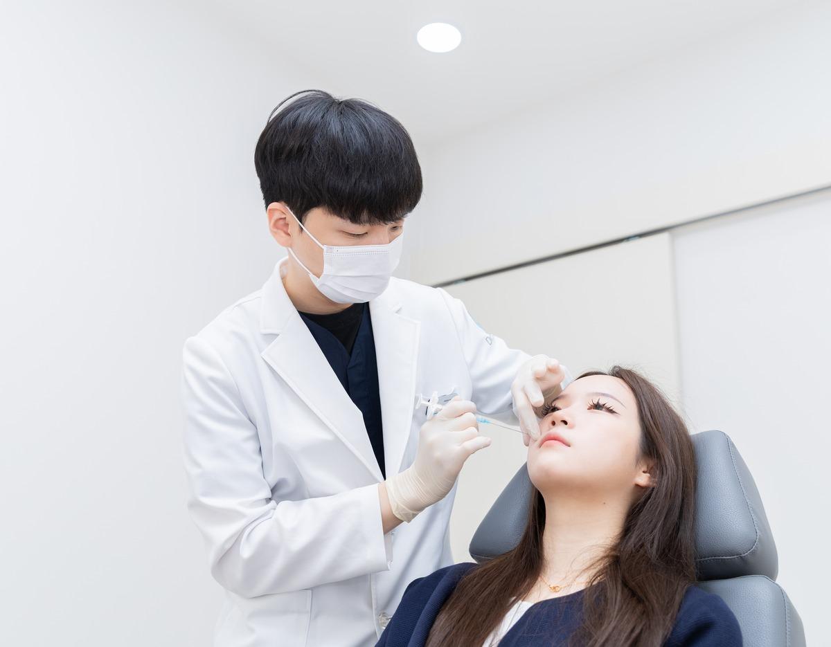 Doctor in white coat administering an injection to a reclining female patient in a bright clinic room