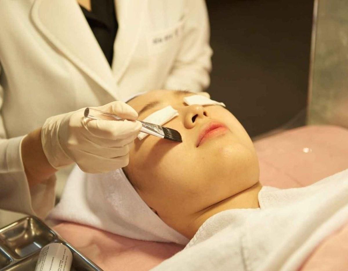 Clinician in white coat wearing gloves performing a facial treatment on a patient lying on a pink bed with towel-covered chest.
