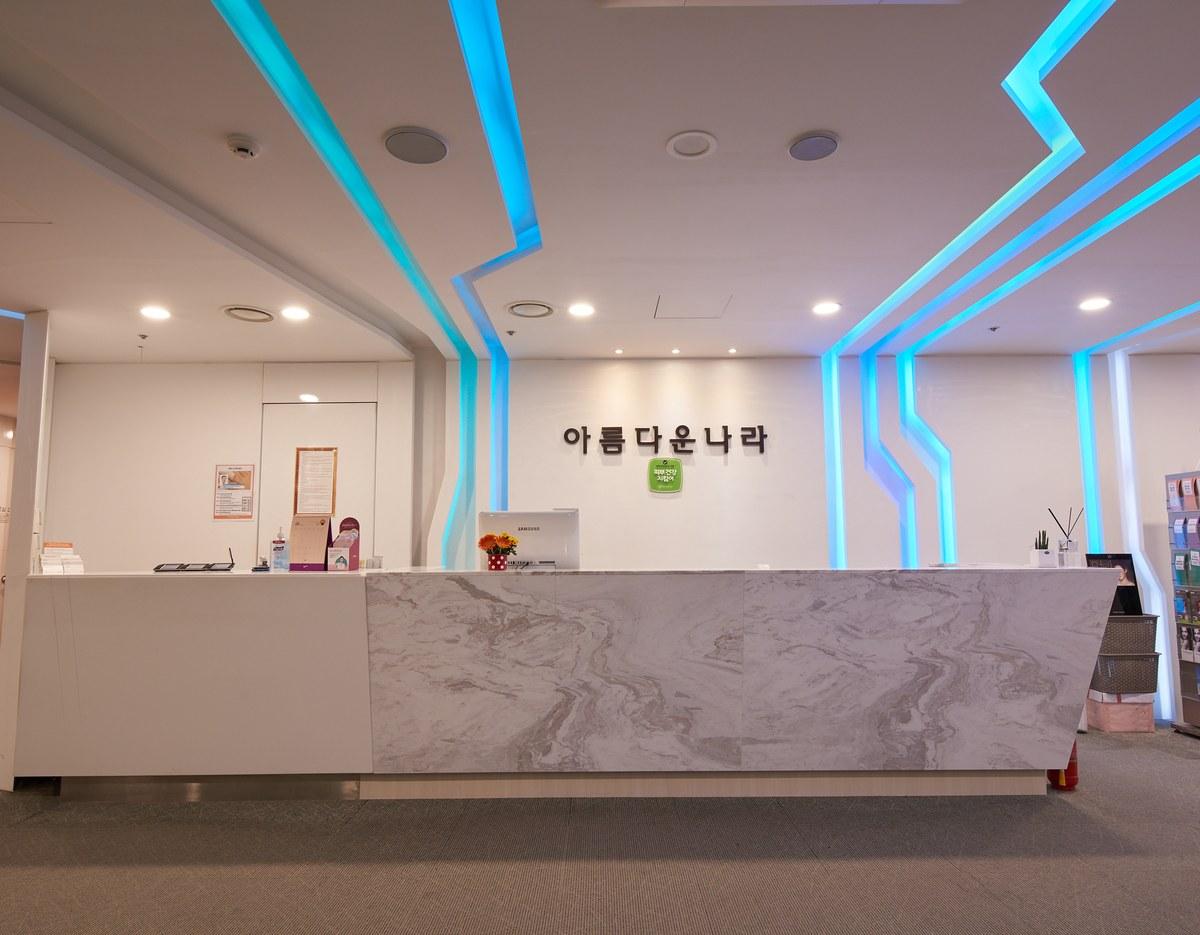 Clinic reception desk with marble-front counter, blue LED ceiling accents and Korean sign "아름다운나라" on white wall.