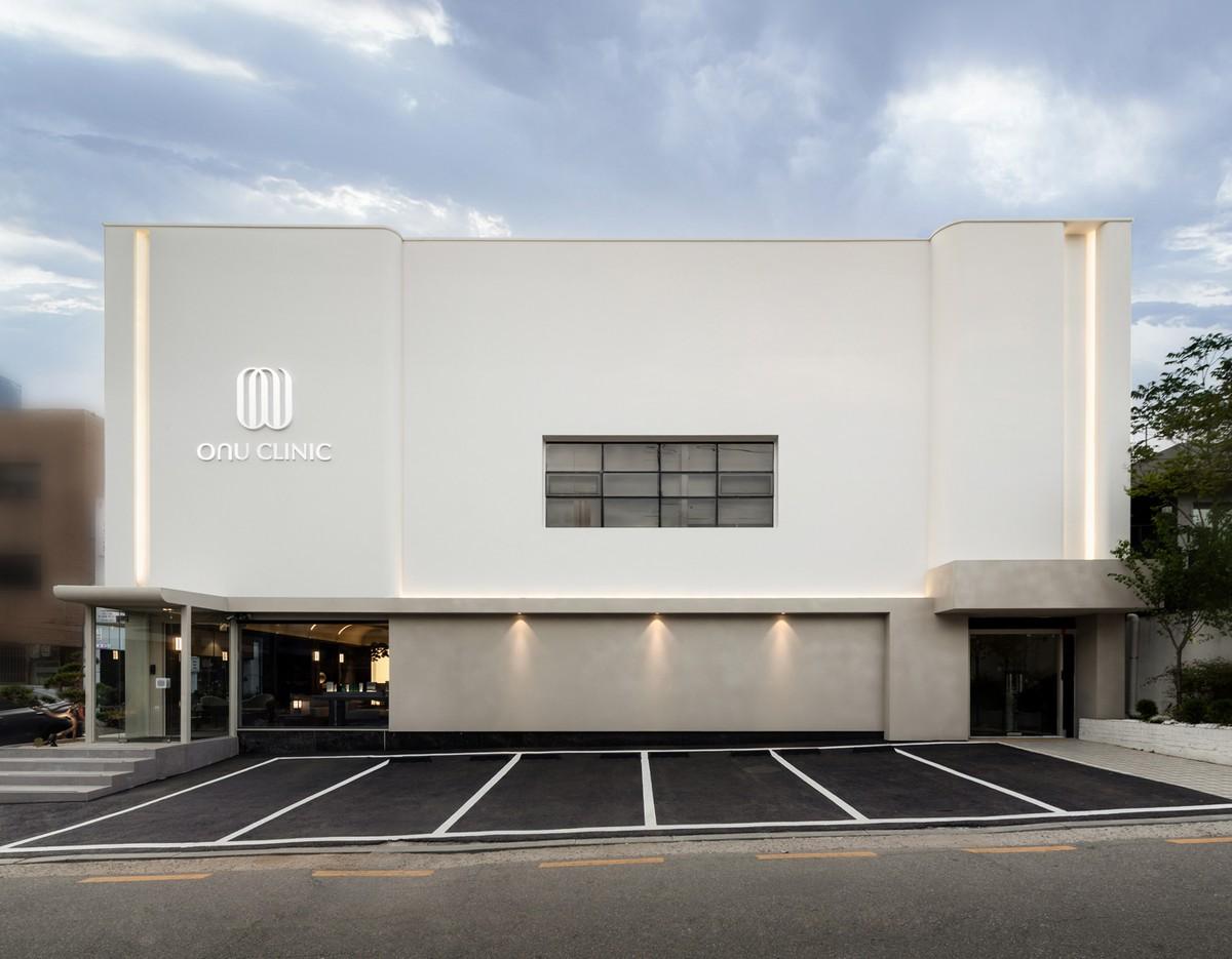Onu Clinic exterior: minimalist two-story white building with logo, glass entrance and empty marked parking spaces under cloudy sky