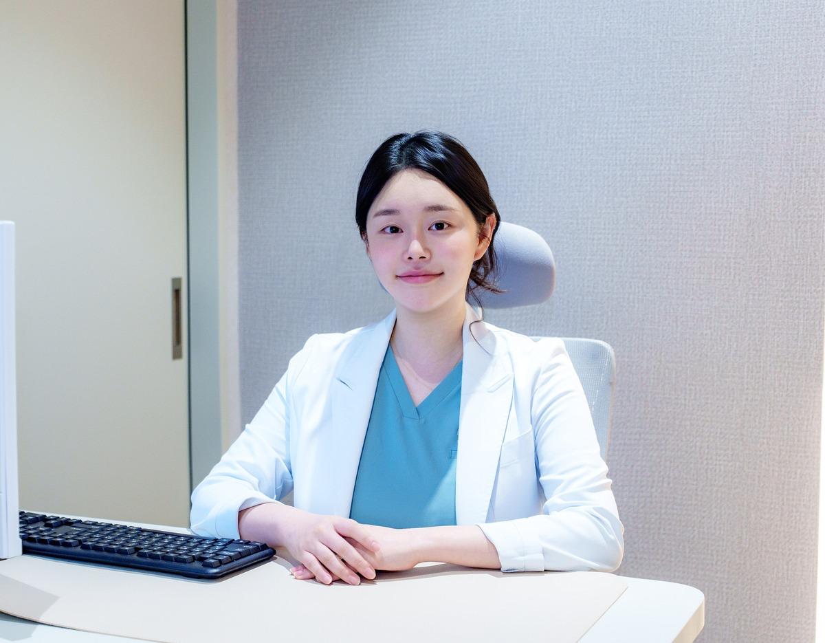 Onu Clinic representative doctor seated at desk in white coat and teal scrubs with hands folded, clinical office background