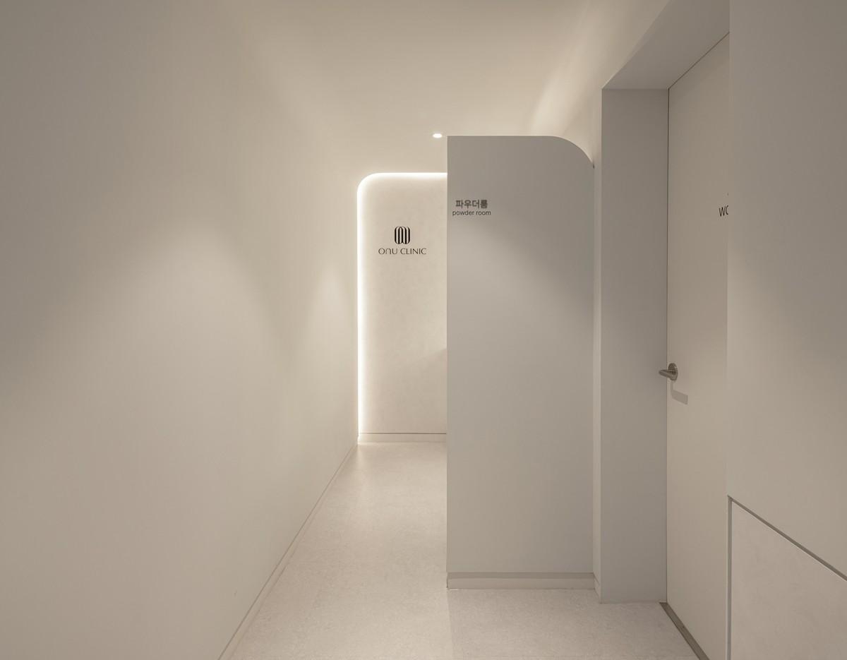 Minimalist pale hallway with curved partition, backlit Onu Clinic logo and signs for powder room and WC.