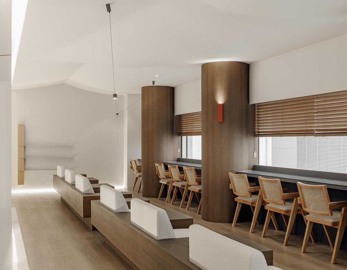 Minimalist clinic waiting area with wooden benches, cushioned backrests, column seating with rattan chairs and long counter windows.