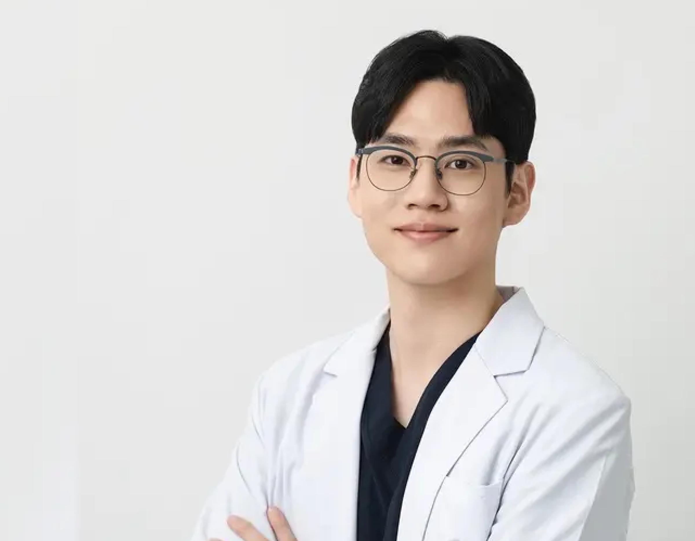 Young male doctor in glasses wearing a white coat and dark scrubs, smiling against a plain light background